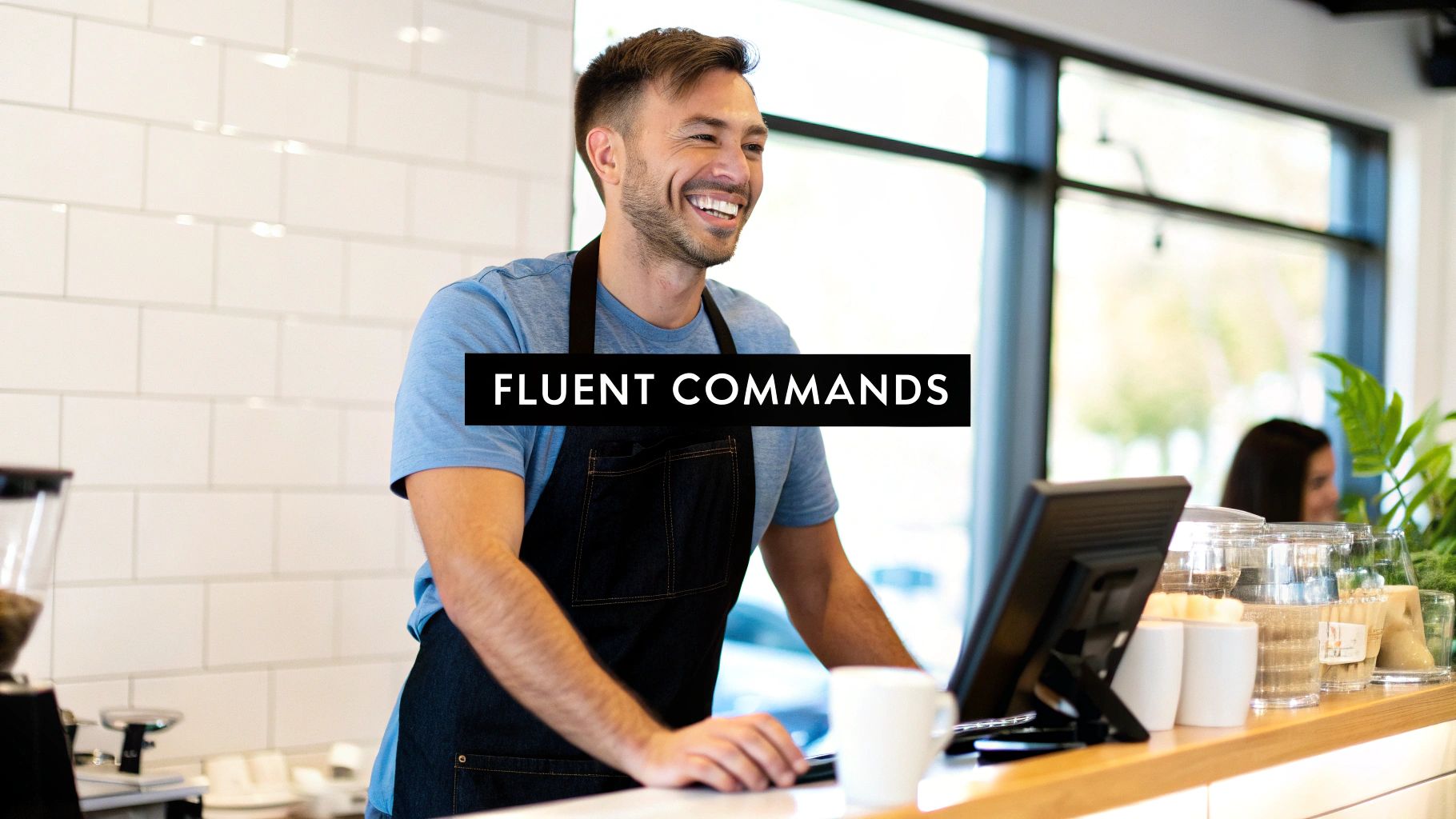 A smiling male barista in a blue shirt and black apron stands behind a cafe counter with a POS system.