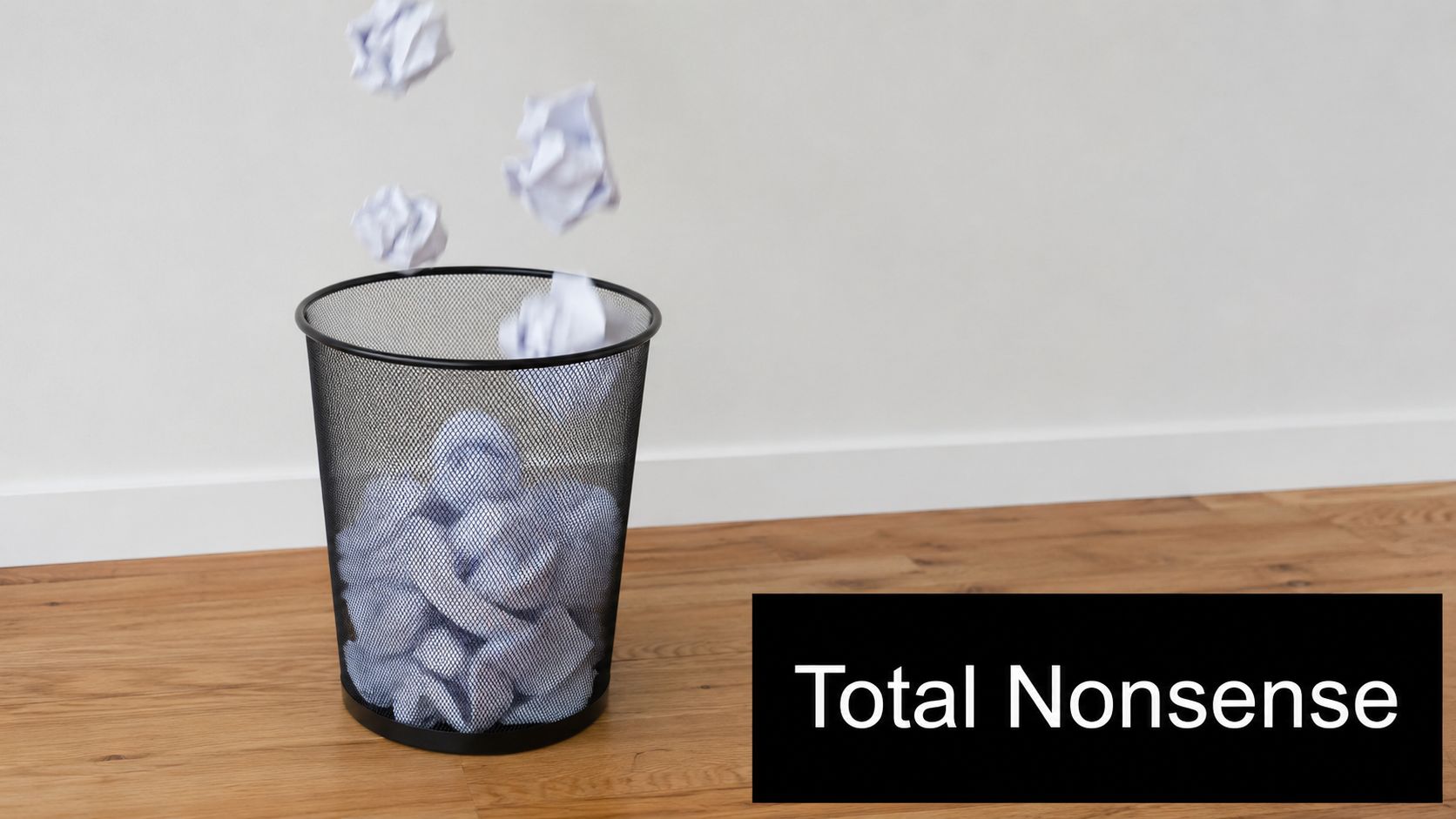 Crumpled paper balls falling into a black wire mesh wastebasket on a wooden floor.