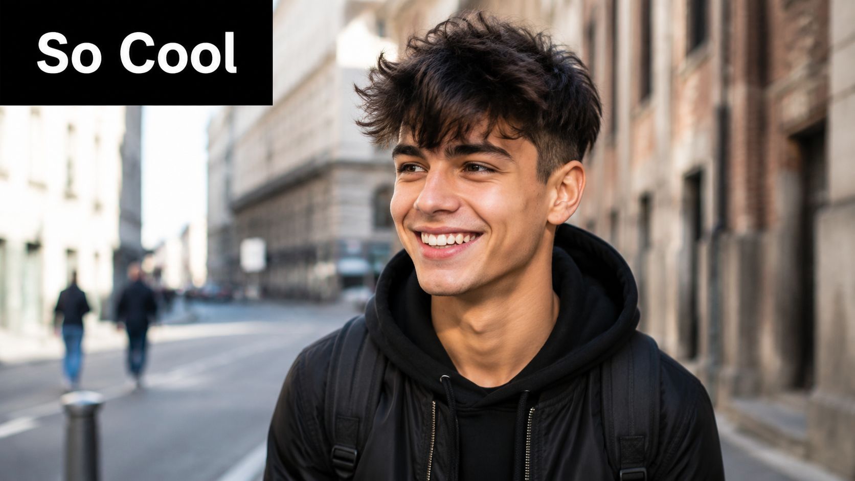 A smiling young man with stylish messy dark hair wearing a black hoodie walking outdoors.