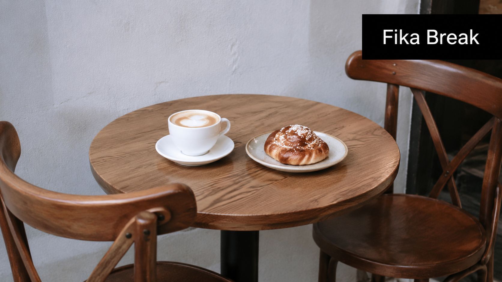 A cup of latte and a traditional cinnamon bun on a wooden table between two chairs.